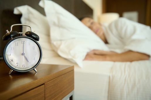 Soft focus on black alarm clock on nightstand next to a blurred bed with sleeping woman. Copy space for advertisement. Healthy sleep and wake up concept