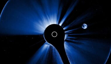 A blue-tinted image of the Sun is partially obscured by a circular black disk in the center, while a small, distant Earth appears on the right side of the image against the blackness of space.