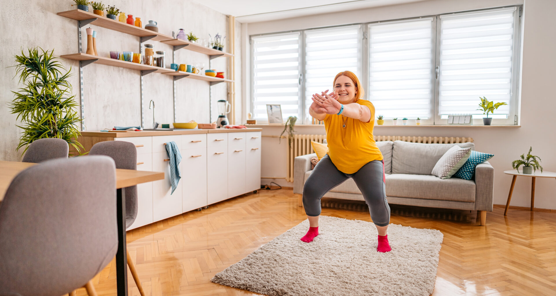 A woman wearing leggings and a t-shirt practices squats at home. She is standing on a rug, behind her we see a couch and to her side there is a kitchen unit with a sink and dishes.