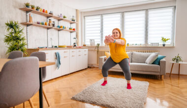 A woman wearing leggings and a t-shirt practices squats at home. She is standing on a rug, behind her we see a couch and to her side there is a kitchen unit with a sink and dishes.