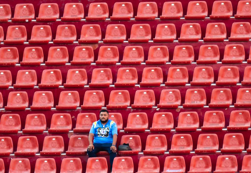 A fan sits alone in a stand at a stadium with rows of empty red seats all around him