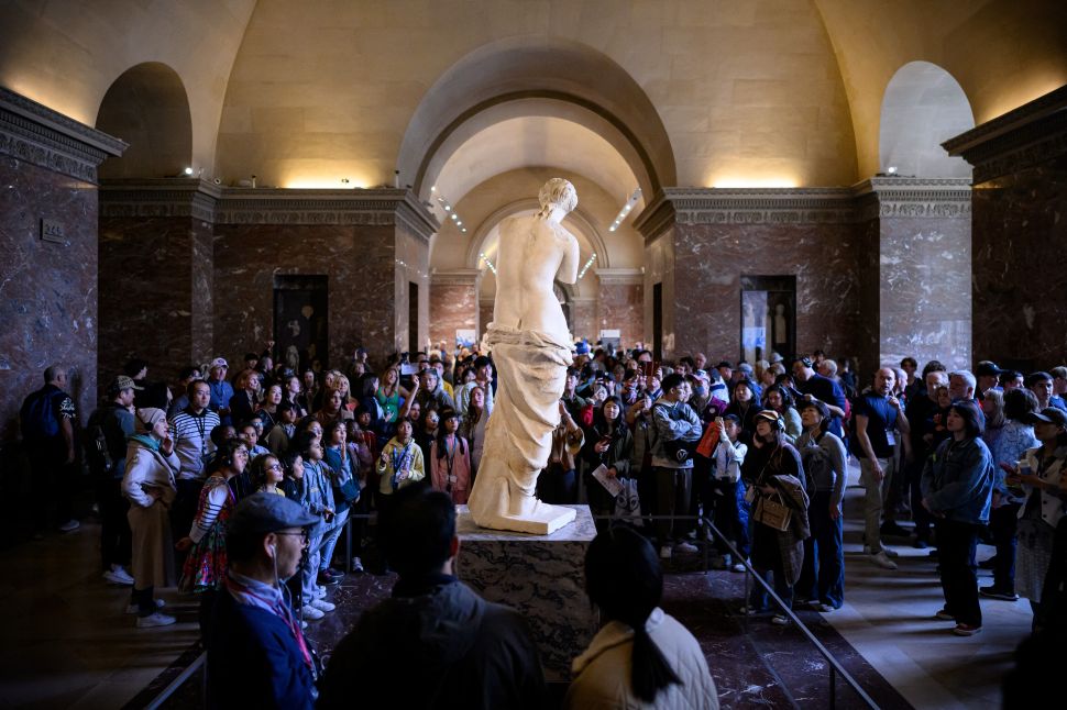 A large crowd of visitors gathers around the Venus de Milo statue inside the Louvre Museum in Paris, viewing and photographing the iconic marble sculpture.
