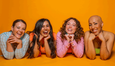 The four members of the girl group lie on the ground, with their faces on their crossed arms. Background is a mustard yellow. All are smiling.