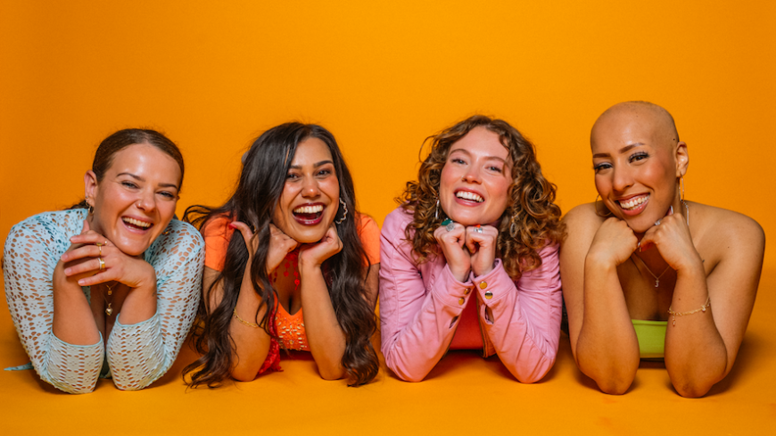 The four members of the girl group lie on the ground, with their faces on their crossed arms. Background is a mustard yellow. All are smiling.