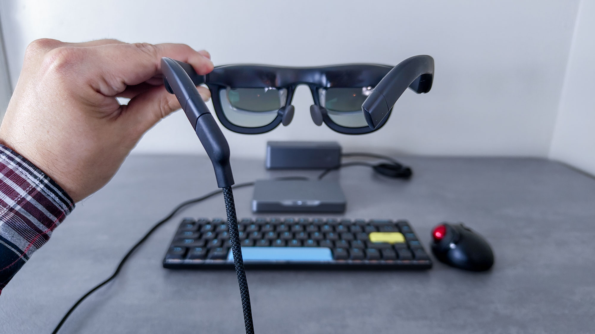 Holding up the Xreal One AR glasses in front of the Khadas Mind 2S, a mechanical keyboard, a trackball mouse and a Ugreen power bank on a desk