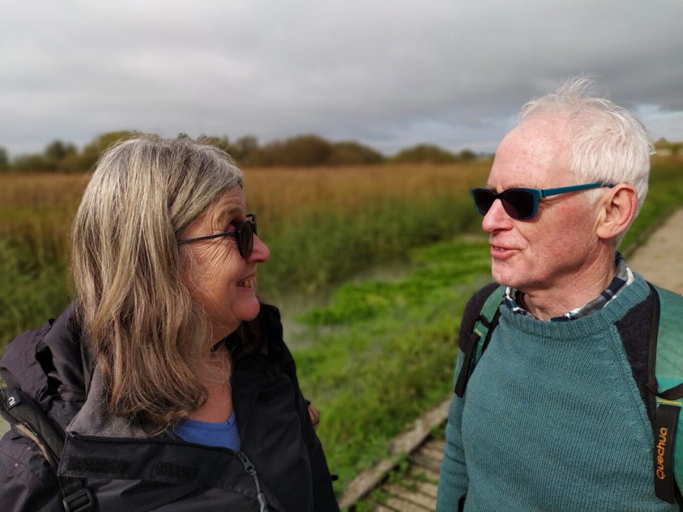 A photo of a man and a woman standing near each other and smiling at each other. They're both wearing dark sunglasses and appear to be in the middle of a field on a hike