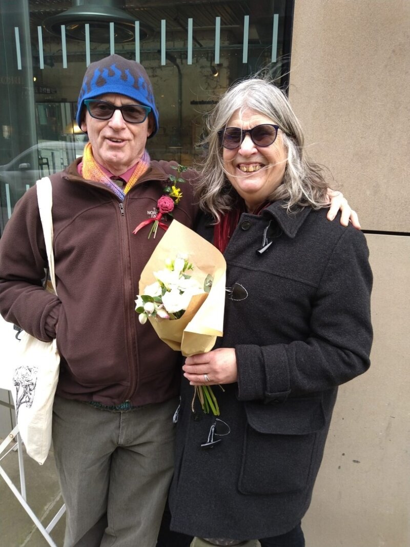 A photo of a man and a woman standing side-by-side, with his arm around her. She's holding a bouquet of flowers and they're both smiling