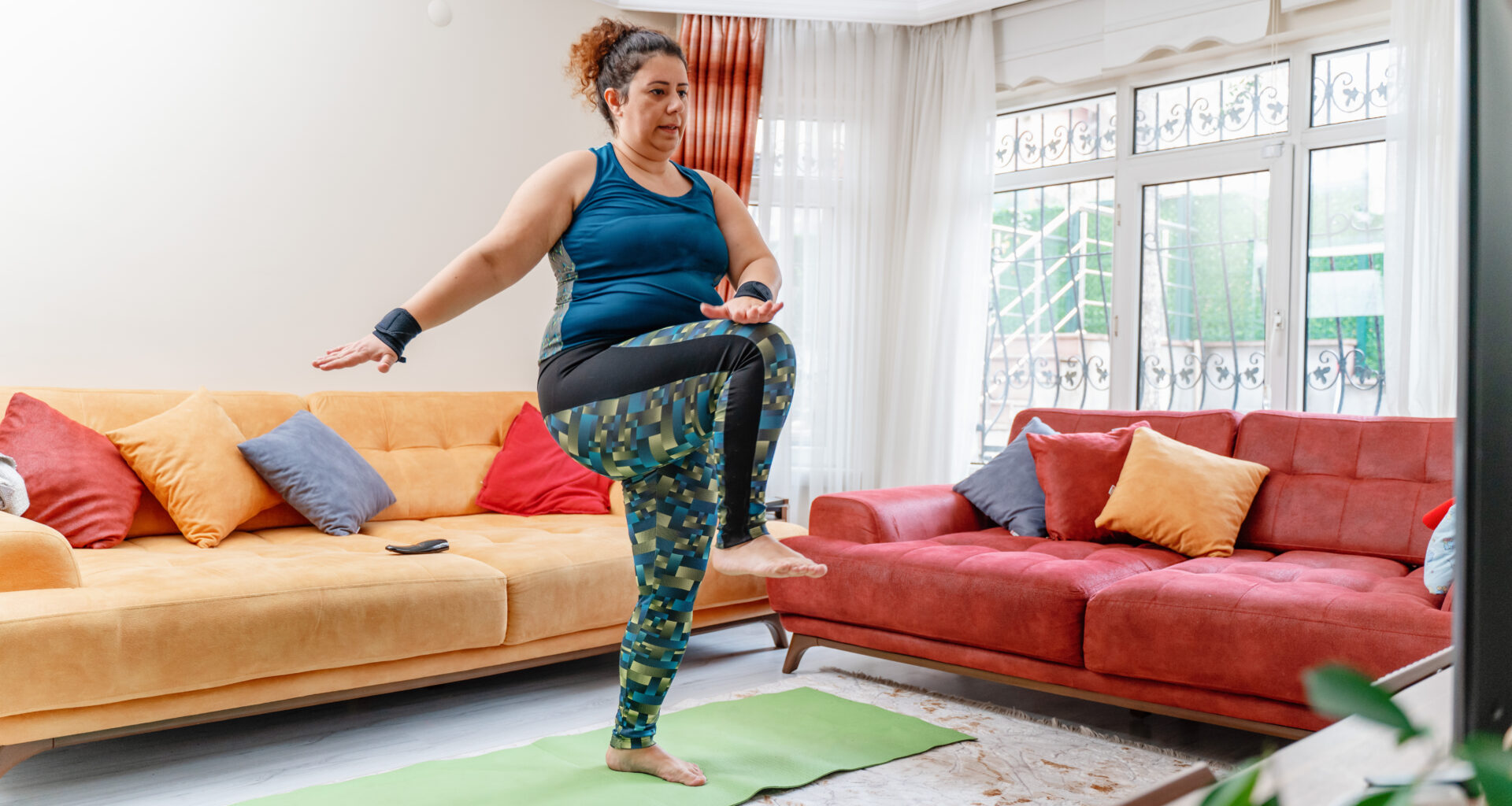 A woman works out at home in leggings and a vest. She is standing on an exercise mat, marching, with her right leg raised and her left hand touching her right knee. Behind we see two couches.