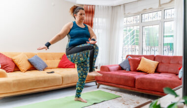 A woman works out at home in leggings and a vest. She is standing on an exercise mat, marching, with her right leg raised and her left hand touching her right knee. Behind we see two couches.