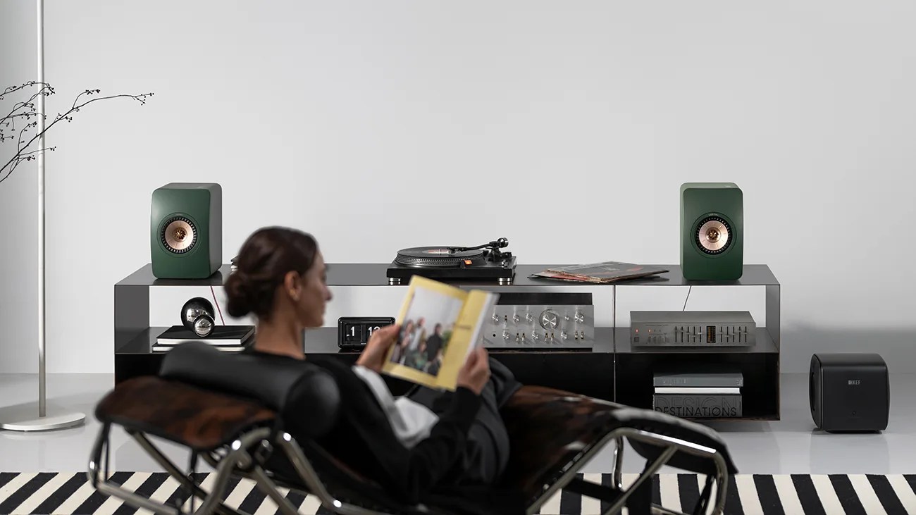 A modern audio setup with two green bookshelf speakers featuring copper-colored cones, a turntable, and audio equipment on a sleek black media console. A black subwoofer is placed on the floor to the right. A person is seated in a black and brown lounge chair in the foreground, reading a book. The room has a minimalist design with a white wall and a black and white striped rug.