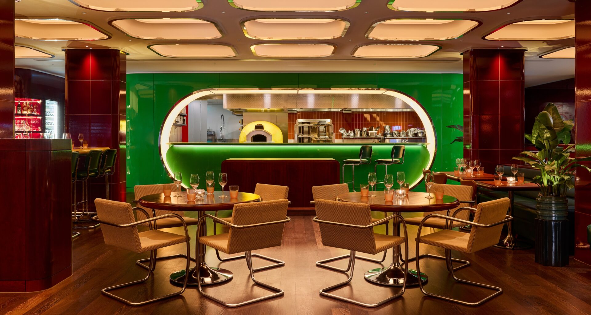 An immersive restaurant with coffered, backlit ceilings, an oblong, lacquered, green kitchen, and leather and chrome chairs boasts cherry plum columns in curved ceramic tiles and a warm atmosphere.