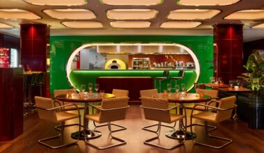 An immersive restaurant with coffered, backlit ceilings, an oblong, lacquered, green kitchen, and leather and chrome chairs boasts cherry plum columns in curved ceramic tiles and a warm atmosphere.