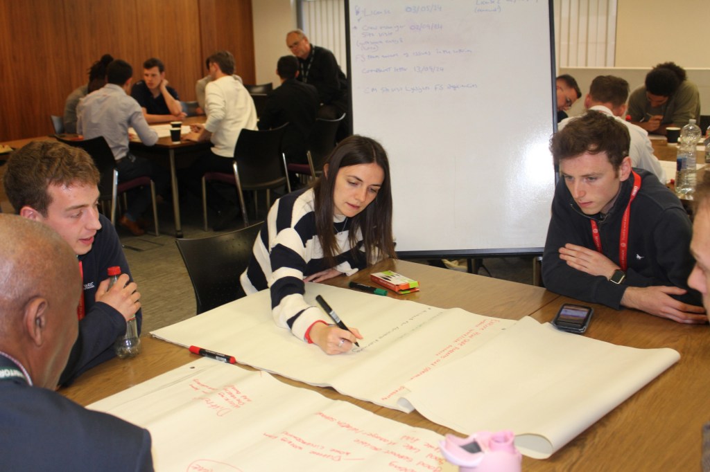 Five people sitting round a table - as a woman in a striped jumper writes on flip chart paper with a marker pen