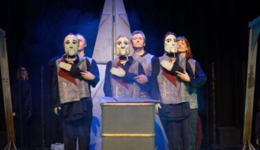 Performers on stage with the puppet masks, stand in the centre, large item at the back behind them