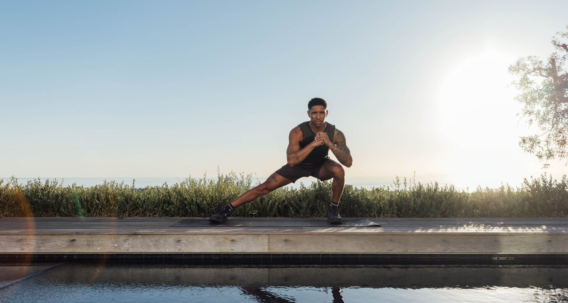Man demonstrates side lunge exercise outside, he is reflected in a body of water in front of you. There is grass and blue sky behind him.