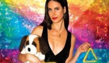 Brooke in a black top and trousers, holding a toy dog, with a rainbow coloured background.