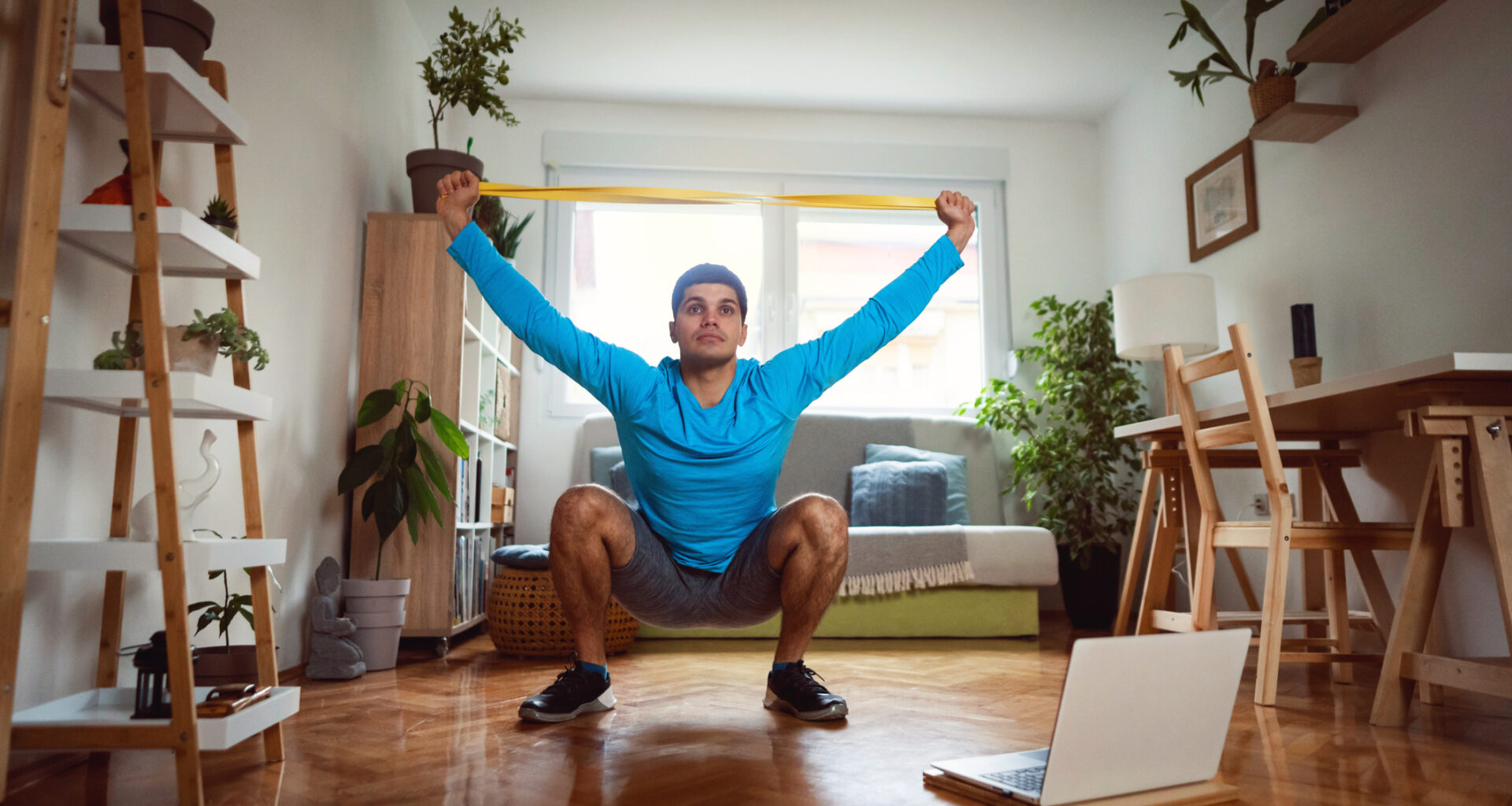Man in sportswear in living room sits in a deep squat holding a resistance band in both hands above his head.