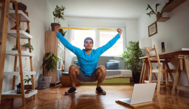 Man in sportswear in living room sits in a deep squat holding a resistance band in both hands above his head.