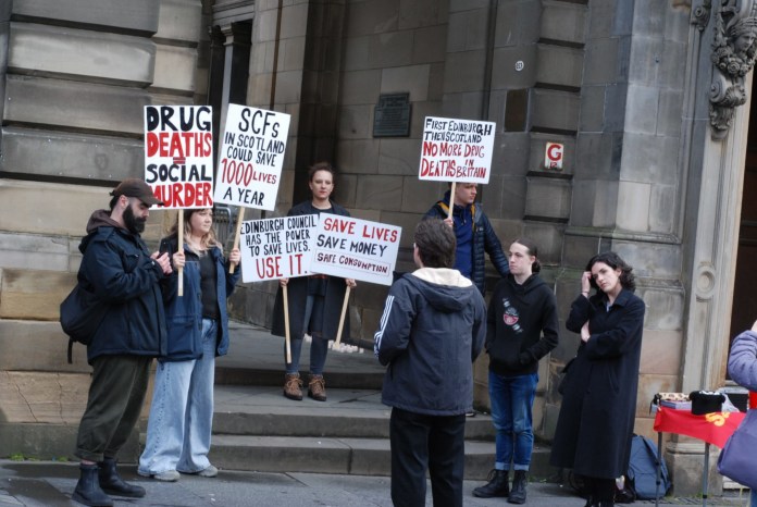 A group of people protesting at the Edinburgh City Chambers. They are holding signs which say phrases such as "Drug deaths = social murder", "SCFs in Scotland could save 1000 lives a year", "Edinburgh Council has the power to save lives. Use it", "Save lives. Save money. Safe consumption." and, "First Edinburgh then Scotland. No more drug deaths in Britain."