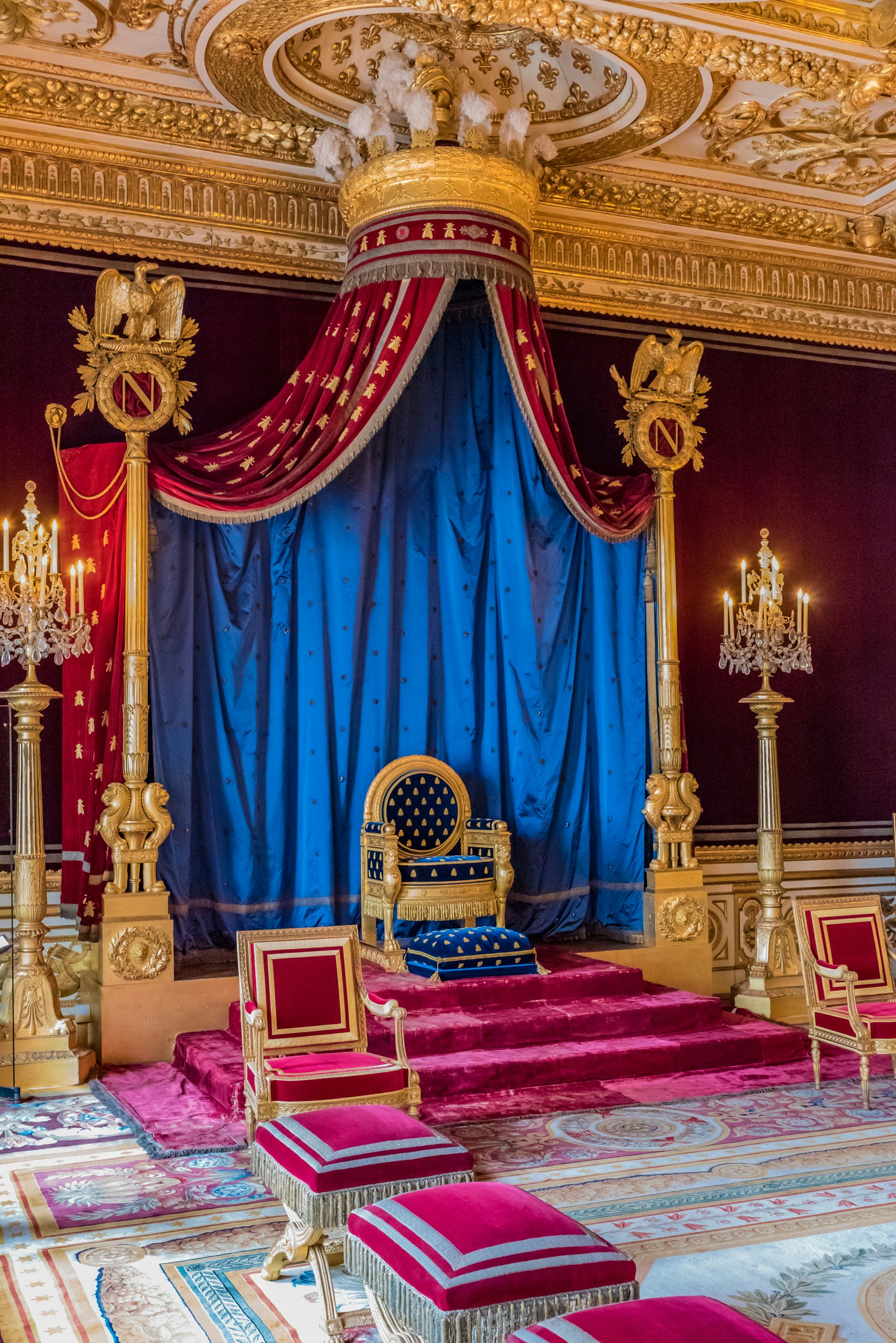 Ornately upholstered plush blue chair with gold trim with a red, blue and gold drape behind it. The letter N is in two staffs on either side of the chair, which is elevated on three red steps.