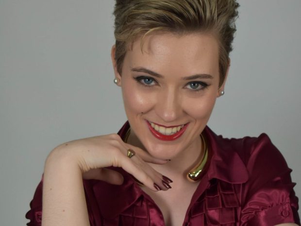 A person with short, styled hair smiles confidently, wearing a burgundy top with ruffled detail and a chunky necklace, against a plain background.