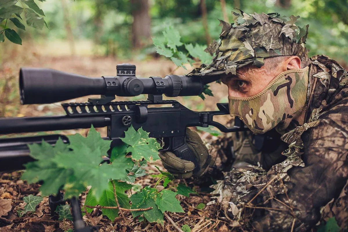 A sniper lies in the foliage wearing a camouflaged outfit, holding a rifle with a scope