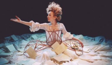 A woman sits on the ground, surrounded by a big white dress. Her hair is piled high and messy and one arm is outstretched.