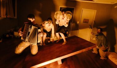 A musican plays; 3 other actors hide behind objects in a dark, shadowy room.