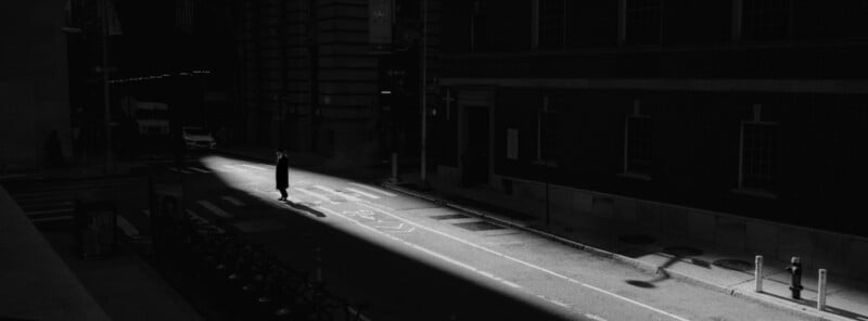 A person in a long coat stands alone in a beam of sunlight on a mostly dark, empty city street, with buildings casting deep shadows around them.
