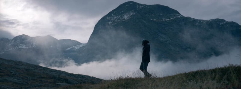 A person stands on a grassy hill in front of a large, misty mountain under a cloudy sky. The scene is serene and dramatic, with low clouds drifting around the mountain peaks.