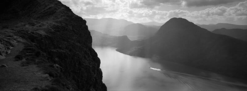 Black and white photo of a lake surrounded by mountains, with sun rays shining through clouds and a small boat creating ripples as it speeds across the water. Rocky terrain is visible on the left side.