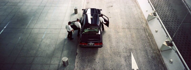 A black car with the rear door open is parked on a street next to a building. Two people in white shirts stand by the car, one by the door, and another by the trunk. The scene is viewed from above.