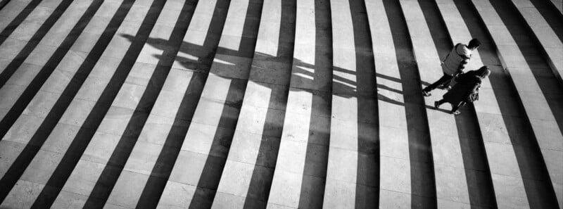 Aerial view of two people standing on wide steps with alternating light and shadow stripes, casting a long shadow as they face each other.