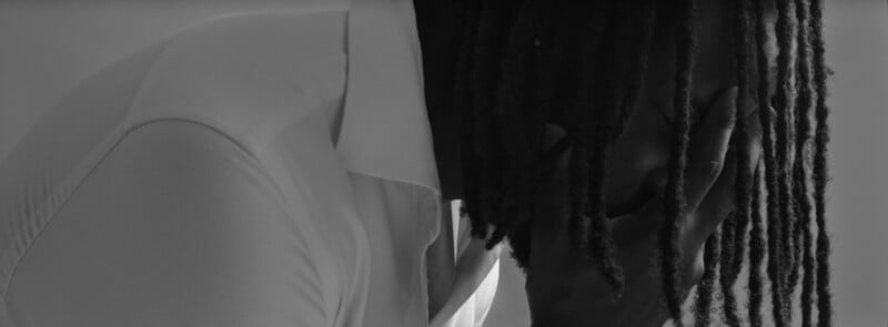 A person wearing a white shirt leans forward, covering their face with one hand. Their long, dark dreadlocks hang down, partially obscuring their face. The image is in black and white, evoking a somber mood.