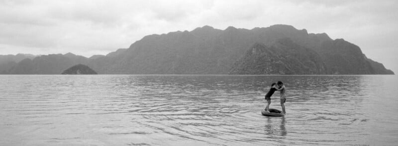 Two people stand closely together on a small raft in calm water, surrounded by ripples, with large, misty mountains in the background under a cloudy sky.