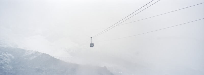 A cable car travels through misty, snowy mountains, suspended on cables above a rocky slope, surrounded by dense fog and limited visibility.