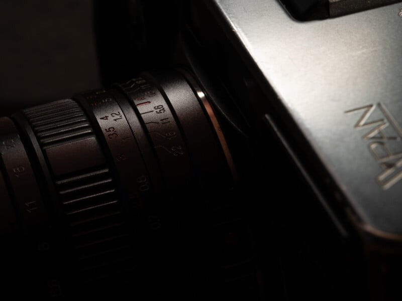 Close-up of a vintage camera lens showing aperture and focus rings, with part of the camera body visible and the word "Japan" engraved on the metallic surface. Lighting is low, creating a moody atmosphere.