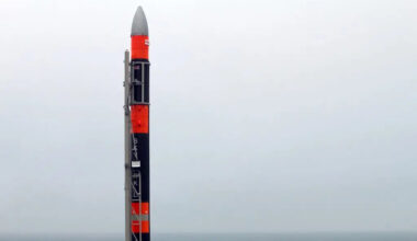 A large rocket with alternating red and black stripes stands on the coast overlooking a cloudy sky.