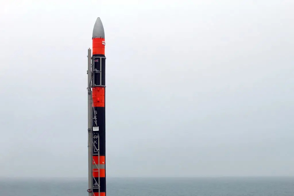 A large rocket with alternating red and black stripes stands on the coast overlooking a cloudy sky.