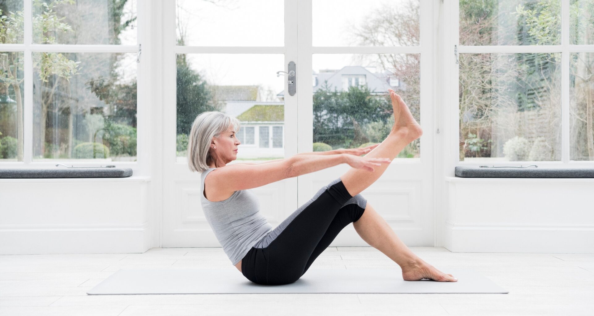 Woman in her 60s with grey bobbed hair stretching leg at home in front of windows overlooking a garden.