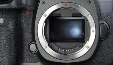 Close-up of a DSLR camera body with no lens attached, revealing the sensor inside a metal mount. The camera grip has a textured finish, conveying durability