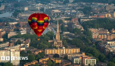 A multi coloured hot air balloon flying over Bristol. The city landscape, consisting of buildings and water, can be seen below.
