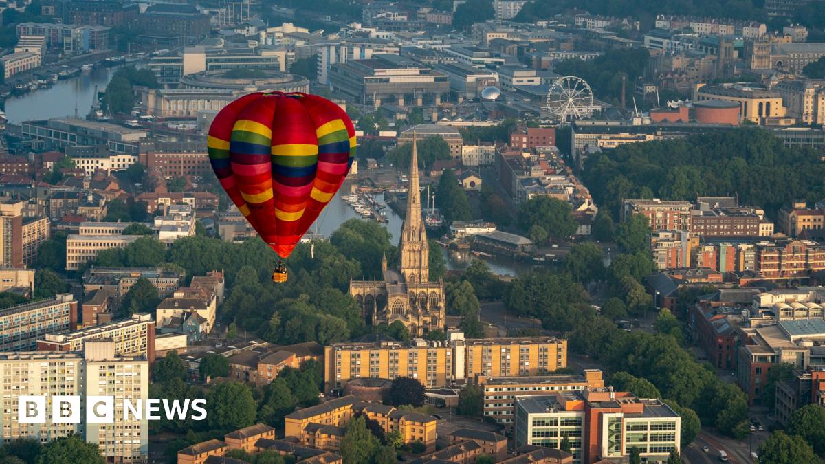 A multi coloured hot air balloon flying over Bristol. The city landscape, consisting of buildings and water, can be seen below.