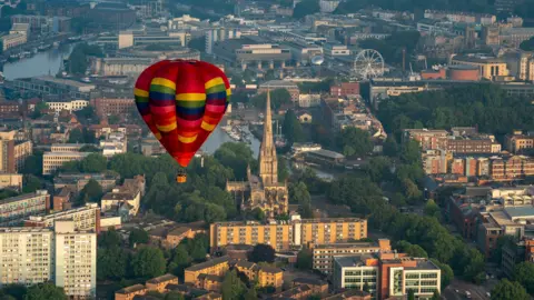 PA Media A multi coloured hot air balloon flying over Bristol. The city landscape, consisting of buildings and water, can be seen below. 