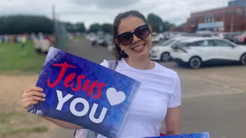 BBC A woman with brown hair and sunglasses smiles at the camera, She is wearing a white t-shirt, and is holding a sign which reads 'Jesus (heart shape) YOU'. There are a number of parked cars behind the woman, as well as a large patch of grass, several trees, and a crowd of people. 