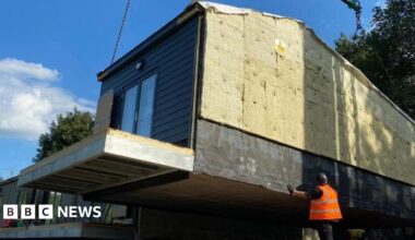 One of the six properties being assembled in a row next to the others. They are built off-site. A crane lifts a near-complete house to its required location whilst a man in an orange high-visibility jacket supervises.