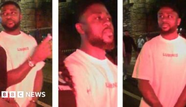 CCTV images of a man on a night out, stood outside on the pavement with other people pictured in the background. He is wearing a white t-shirt and has a watch on. He also has a black beard and black hair.