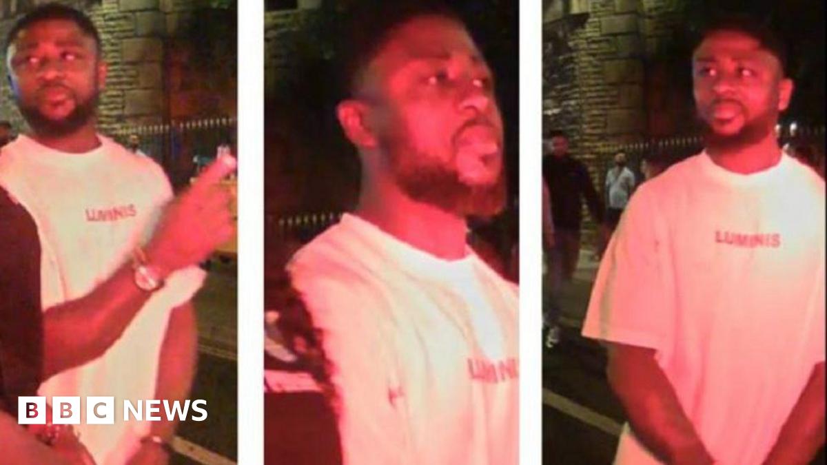 CCTV images of a man on a night out, stood outside on the pavement with other people pictured in the background. He is wearing a white t-shirt and has a watch on. He also has a black beard and black hair.
