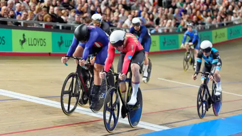 SWPIX Ioan leading a tight pack of cyclists at the velodrome. They are all wearing black and white helmets and cycling Lycra, Ioan wears a red top and black shorts. A crowd can be seen in the background. 