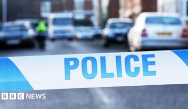 A close up shot of blue and white police tape. It is striped with "police" written on it in blue capitals. In the background there are a number of cars on a residential street, although it is blurred quite heavily.